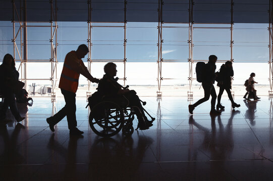 Silhouette Of man Walking With Disabled Person In Wheelchair Airport. People going on flight, large windows on background