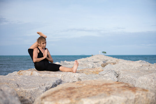 Elderly woman sitting on stone sea  beach holding her leg behind head doing yoga exercises and stretching