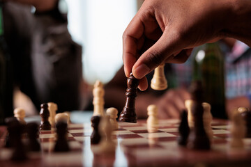 Close up of hand moving chess piece on chessboard while sitting at table. Person playing strategy boardgame with friends while sitting at home in living room with snacks and beverages. - obrazy, fototapety, plakaty