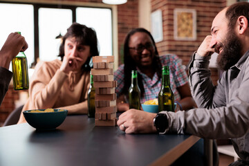Multiethnic happy friends sitting at table in living room while playing society games together. Diverse people having fun by enjoying snacks, beer and fun leisure time activities at home.