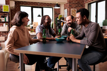 Multicultural group of people sitting at table in living room while laughing and talking together. Multiethnic friends having fun while enjoying snacks and beverages while sitting at table.