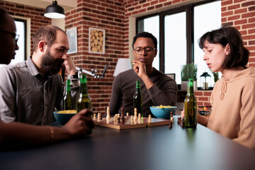 Pensive african american man thinking about next chess move while sitting at table. Focused confident person sitting at home in living room with multiethnic friends while playing strategy boardgames.