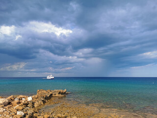 Fototapeta na wymiar The stone coast of the Mediterranean Sea against the backdrop of the sea with a walking white ship and a dramatic sky.
