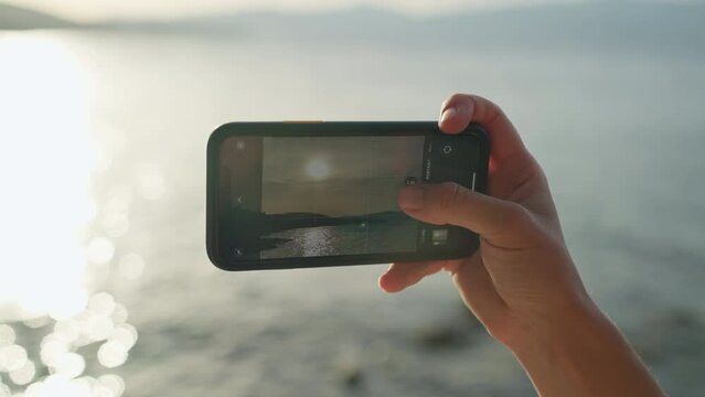 Close-up of a phone in the hands of a woman making a photo at sunset. Beautiful seascape with waves and happy woman on vacation making photo in memory.