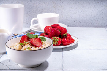Rice porridge or pudding with fresh strawberry and nuts for breakfast, with plate of strawberries, white tiled background copy space