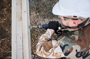 Man building frame house. Male worker drilling hole by electric drill in wooden frame of future house. Builder wearing white helmet and protective goggles. Carpentry concept.