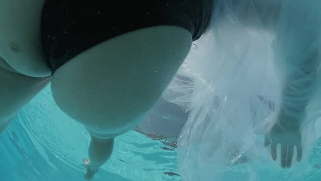 Young woman dressed in a black swimsuit and a white sheer long cape swims in the outdoor swimming pool decorated with blue tiles.
