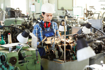 Serious concentrated female factory worker in hardhat standing at machine and processing watch...