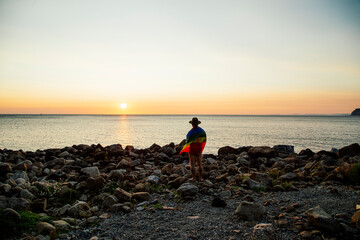 Man with hat and lgtbi flag standing naked on the beach contemplating the sunset