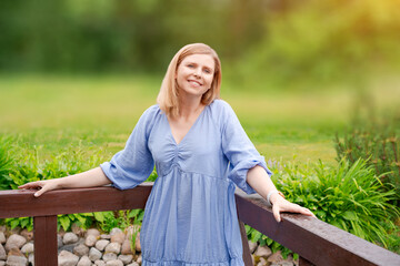 Gorgeous young blond woman in stylish blue dress happy in garden conversations, stands smiling leaning against a wooden fence smiling. Outdoor shot in the park during summer daytime - Powered by Adobe