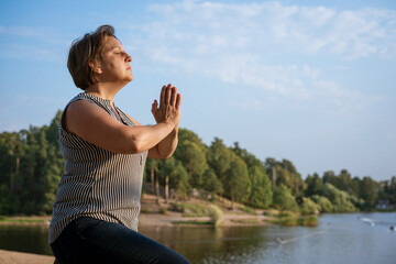 Naklejka na ściany i meble Lonely woman meditates sitting on shore lake with her palms clasped in front her or prays. The concept of relaxation and spiritual balance and connection with nature