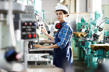 Serious thoughtful woman engineer in uniform standing at industrial machine and producing small gear for wristwatch, she working at production line