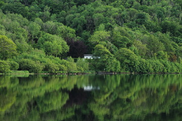 Cottage house nestled amongst greenery of trees in forest reflected in still waters of Lough Gill, situated in rural County Leitrim, Ireland
