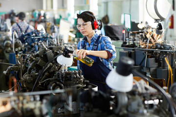 Serious concentrated lady engineer in ear protectors and protective goggles pouring lubricant for machine while working at factory