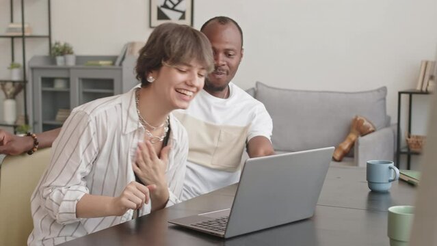 Medium shot of young happy multiethnic couple watching funny dancing videos on laptop and repeating movements, having fun while spending leisure time together at home