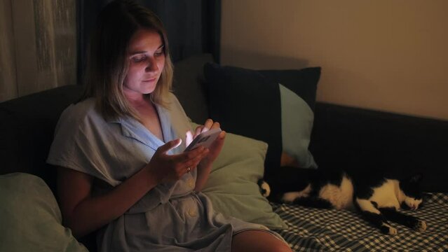 Young woman sit on sofa with cat at home, using smartphone app, spend time in social media, ordering food, sharing news with friends.