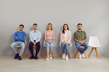 Diverse multiracial millennial young people sit on chairs in line waiting for interview in office. International work candidates or applicant in queue apply for job vacancy. Recruitment concept.