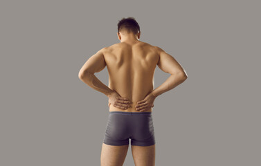 Fototapeta na wymiar Naked man in underwear on grey background suffer from backache or spasm. Pain in lower spine. Guy struggle with painful feeling in back, have kidney stones or inflammation. Healthcare concept.