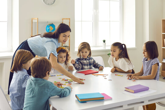 Preschool teacher helps a group of children put together a puzzle. A woman teacher helps children in a preschool group. A group of children at the table collecting a puzzle