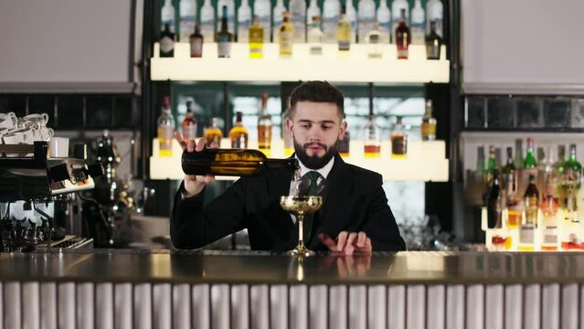 Skillful bartender pouring alcoholic drink from bottle in golden antique goblet while standing behind counter of luxury restaurant. Concept of people, service and nightlife.