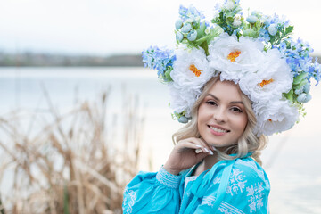 Beautiful young Ukrainian woman dressed in a blue embroidered dress with a large beautiful floral wreath on her head smiling, looking at the camera