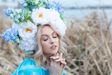 A beautiful young Ukrainian woman dressed in a blue embroidered dress with a large beautiful flower wreath on her head closed her eyes and folded her arms in prayer