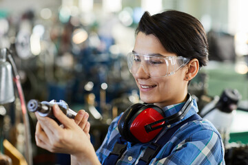 Smiling satisfied young woman in transparent goggles wearing ear protectors examining metal drill bit while choosing detail for metalworking - obrazy, fototapety, plakaty