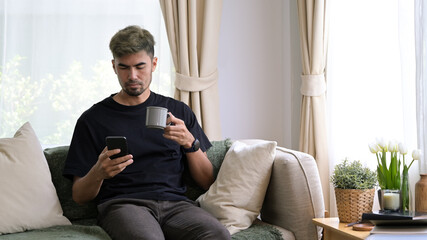 Young man sitting on comfortable sofa and using smart phone, enjoying leisure weekend time at home