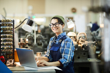 Smiling attractive young female worker of watch plant standing at desk with metal chips and using laptop while looking at camera