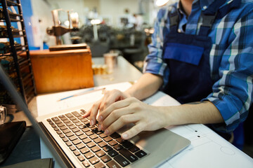 Close-up of unrecognizable woman engineer standing at desk with project plan and using laptop while...