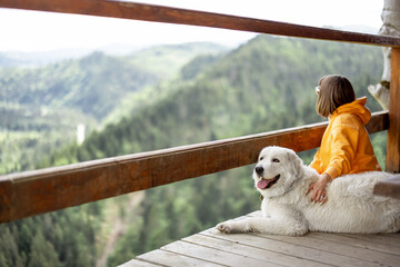 Young person in orange sport suit enjoys great mountain landscape while sitting with her dog on a wooden terrace. Concept of escape and solitude in nature and traveling with pets - Powered by Adobe