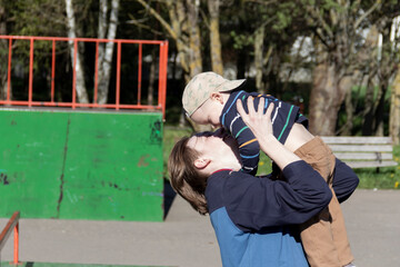 Naklejka premium A happy older brother, a teenager, holds his little, cheerful brother in his arms on a walk in the park, in the yard.