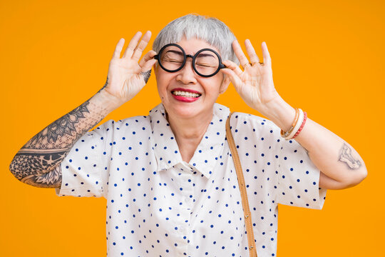 Portrait of attractive funky asian tattoo cheerful grey-haired elder old senior woman touching spectacles isolated over yellow pastel background color,surprise and over exited feeling expression