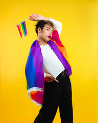 young man with pride movement LGBT Rainbow flag on shoulder isolated yellow background. Man with a gay pride flag looking at camera.