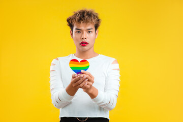 Hopeful Young asian gay man holding rainbow paper cut heart in concept of lgbtq and lgbtq+ posing isolated yellow background.