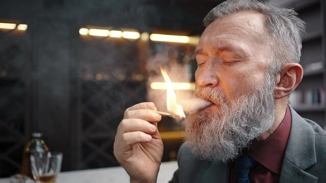 Zoom out portrait of bearded mature businessman smoking cuban cigar with burning match, sitting at luxury cabinet