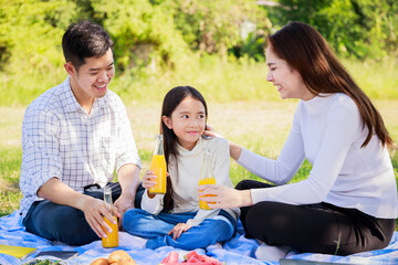 Happy family picnic. Asian parents, little girl drinking orange juice and enjoyed picnic together