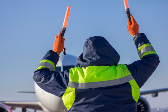 An Airliner Is Taxiing Into A Parking Lot At An Airport, An Air Marshall With Batons Indicates A Parking Spot, An Aeronautical Engineer Helps Pilots Park The Plane