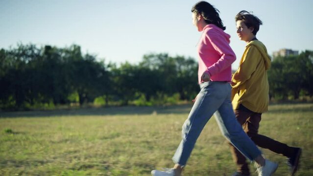 Tracking shot side view of carefree happy teenage boyfriend and girlfriend running in sunshine on green meadow. Wide shot excited loving teen Caucasian couple having fun enjoying dating