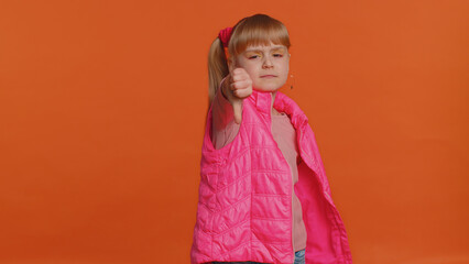 Upset unhappy toddler girl in pink blouse, vest showing thumbs down sign gesture, expressing discontent, disapproval, dissatisfied, dislike. Young little child. Indoor studio shot on orange background