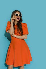 Beautiful young woman in orange cocktail dress and sunglasses is looking away and laughing. - 508405219