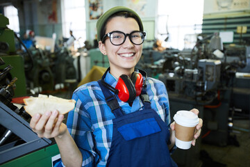 Happy excited lady worker in glasses eating sandwich and drinking coffee from disposable cup while...
