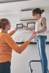 Fototapeta premium Mother and daughter on the ladder hanging pictures and photos on the shelf and wall at home.