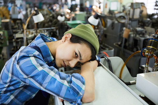 Upset frowning female factory worker in beanie hat tired from work leaning on equipment in industrial shop