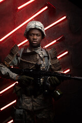 Shot of black soldier dressed in camouflage armor posing against red background with neon.