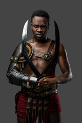 Fototapeta na wymiar Portrait of serious african arena fighter dressed in armor holding twin swords against grey background.