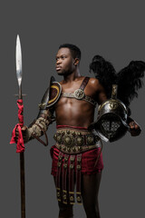 Portrait of muscular african gladiator holding feathered helmet and spear isolated on gray...