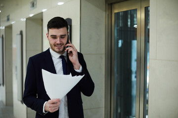 Cheerful confident handsome male manager in black suit walking by elevators and talking to business partner on phone while reading papers