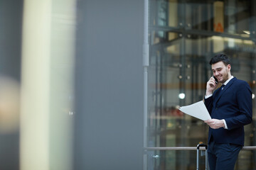 Positive handsome young male manager in suit talking by mobile phone and examining document while waiting for elevator, he has arrived in office of business partner