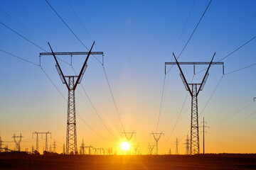 High voltage electricity towers in field at sunset and clear blue sky. Dark silhouettes of...